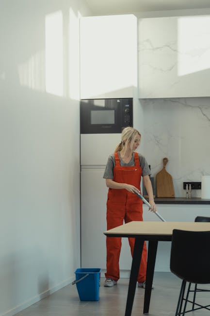 A woman wearing a grey T-shirt and orange overalls is cleaning a light-colored wooden table in a modern kitchen. She is using a mop or cleaning cloth, standing next to a small blue bucket on the floor. The kitchen features a white wall with a built-in microwave and oven, stainless steel stovetop, and minimalist decor with white marble countertops and backsplash. Natural light is illuminating the space, highlighting the clean, polished surfaces. The setting emphasizes surface cleaning and sanitisation as part of professional domestic cleaning services by Elephant and Castle Cleaners in the context of end of tenancy cleaning in Elephant and Castle.