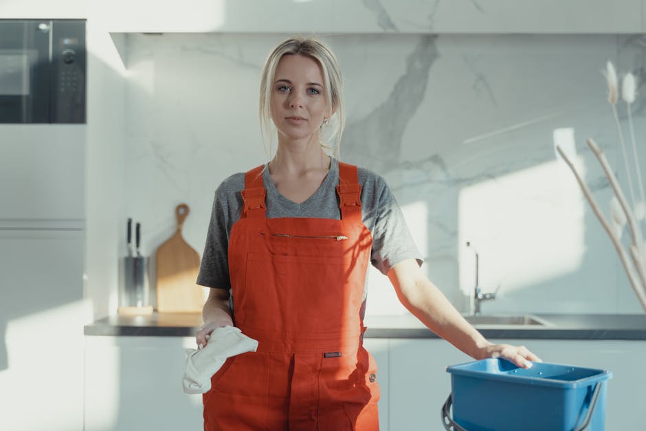 A woman wearing a grey T-shirt and orange overalls is cleaning a light-colored wooden table in a modern kitchen. She is using a mop or cleaning cloth, standing next to a small blue bucket on the floor. The kitchen features a white wall with a built-in microwave and oven, stainless steel stovetop, and minimalist decor with white marble countertops and backsplash. Natural light is illuminating the space, highlighting the clean, polished surfaces. The setting emphasizes surface cleaning and sanitisation as part of professional domestic cleaning services by Elephant and Castle Cleaners in the context of end of tenancy cleaning in Elephant and Castle.