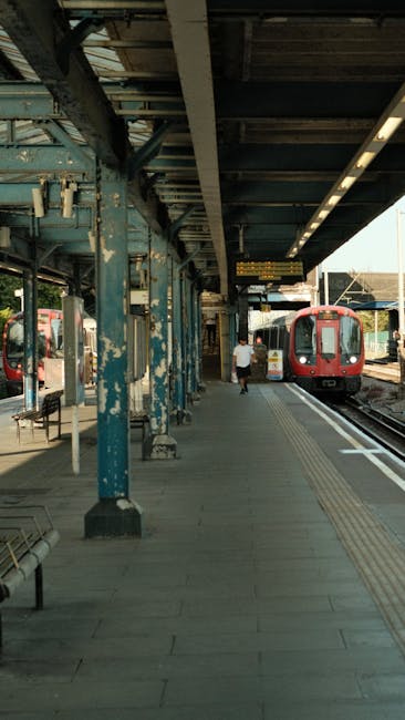 The image shows the platform at Elephant and Castle station, featuring a weathered blue metal canopy with peeling paint and support columns along the edge of the platform. A red train is arriving or departing on the right side, with some seats visible beneath the canopy on the left. The platform surface appears clean and dry, with tactile paving strips along the edge for safety. A passenger in casual clothing is walking towards the train, and the station is illuminated by natural light, creating a bright and clear atmosphere. This setting illustrates the importance of regular surface cleaning and maintenance, with Elephant and Castle Cleaners providing professional sanitisation and deep cleaning services to ensure hygiene and safety in busy transportation hubs.