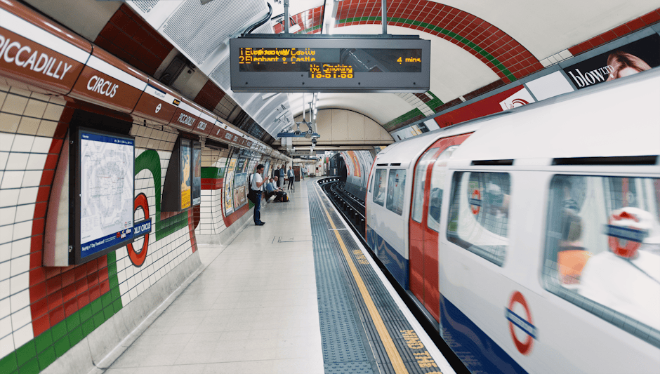 Interior view of Elephant and Castle underground station platform showing a white and red London Underground train arriving at the platform. The platform has tiled walls with green, white, and red accents, digital departure boards overhead displaying travel information, and several passengers waiting, some standing and some seated. The clean tiled floor is well-maintained and free of dust or debris. The lighting is bright and evenly distributed, highlighting the shiny surfaces of the tiles and train. Visible tools include digital screens and seating, with an emphasis on a tidy, hygienic environment. Elephant and Castle Cleaners offers surface cleaning and deep sanitisation services to maintain such high levels of cleanliness in public spaces, aligning with their same-day emergency cleaning service as detailed on their website.