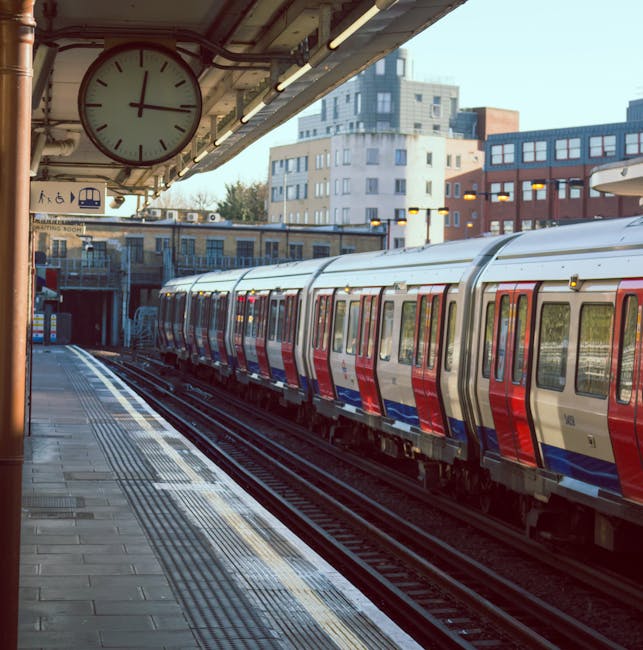 The image shows the platform at Elephant and Castle station, featuring a weathered blue metal canopy with peeling paint and support columns along the edge of the platform. A red train is arriving or departing on the right side, with some seats visible beneath the canopy on the left. The platform surface appears clean and dry, with tactile paving strips along the edge for safety. A passenger in casual clothing is walking towards the train, and the station is illuminated by natural light, creating a bright and clear atmosphere. This setting illustrates the importance of regular surface cleaning and maintenance, with Elephant and Castle Cleaners providing professional sanitisation and deep cleaning services to ensure hygiene and safety in busy transportation hubs.