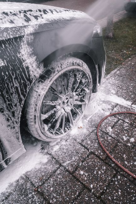 Close-up view of a black car being thoroughly cleaned with soap and water on the front tire and wheel arch, which are covered in soap suds and foam. The car is positioned on a wet, concrete driveway, 